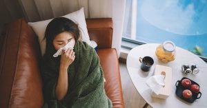 Sick woman lying on sofa in living room covered with blanket and wiping nose with tissue paper.