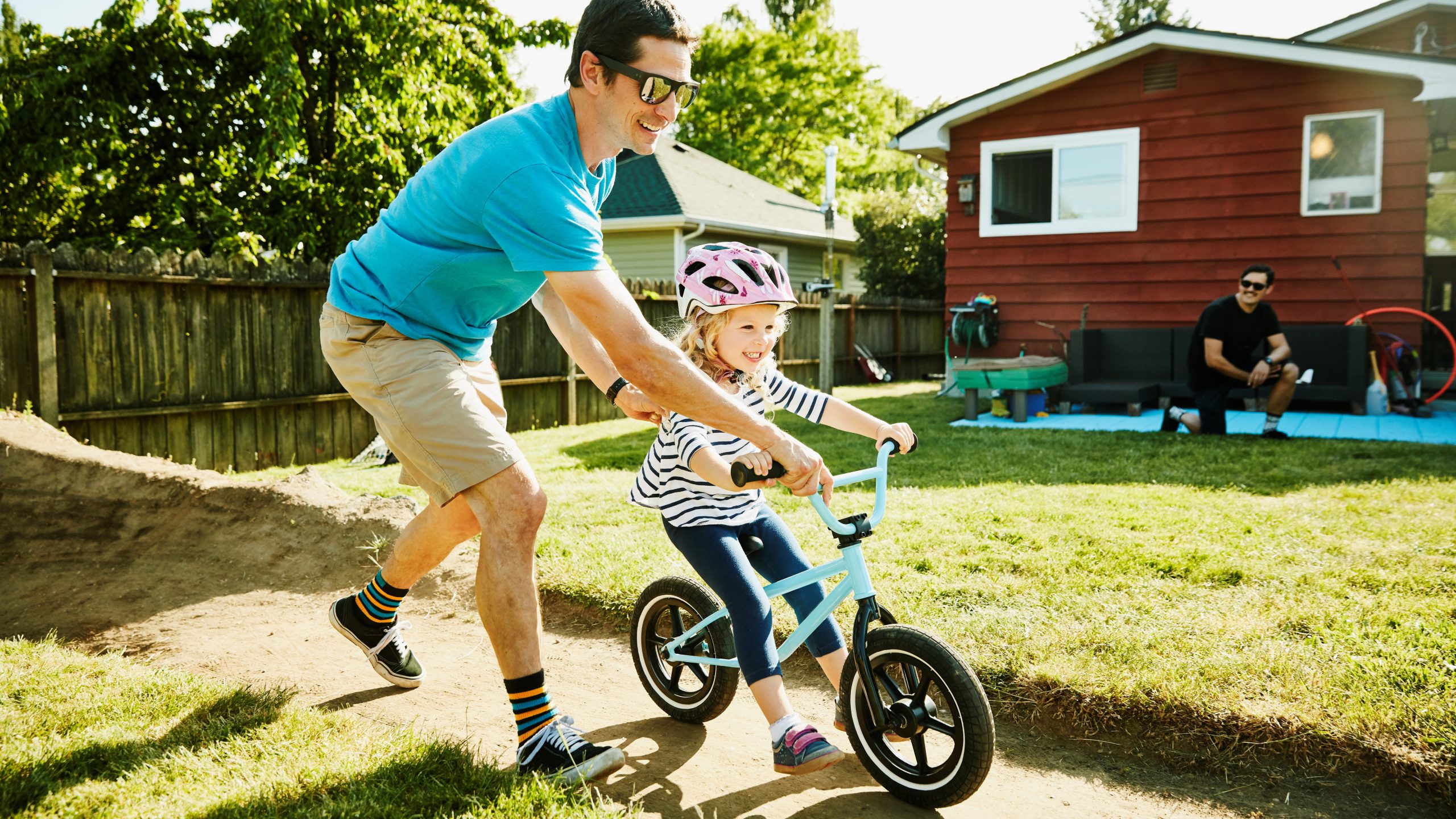 Smiling father pushing young daughter on bike on dirt track in backyard on summer afternoon.