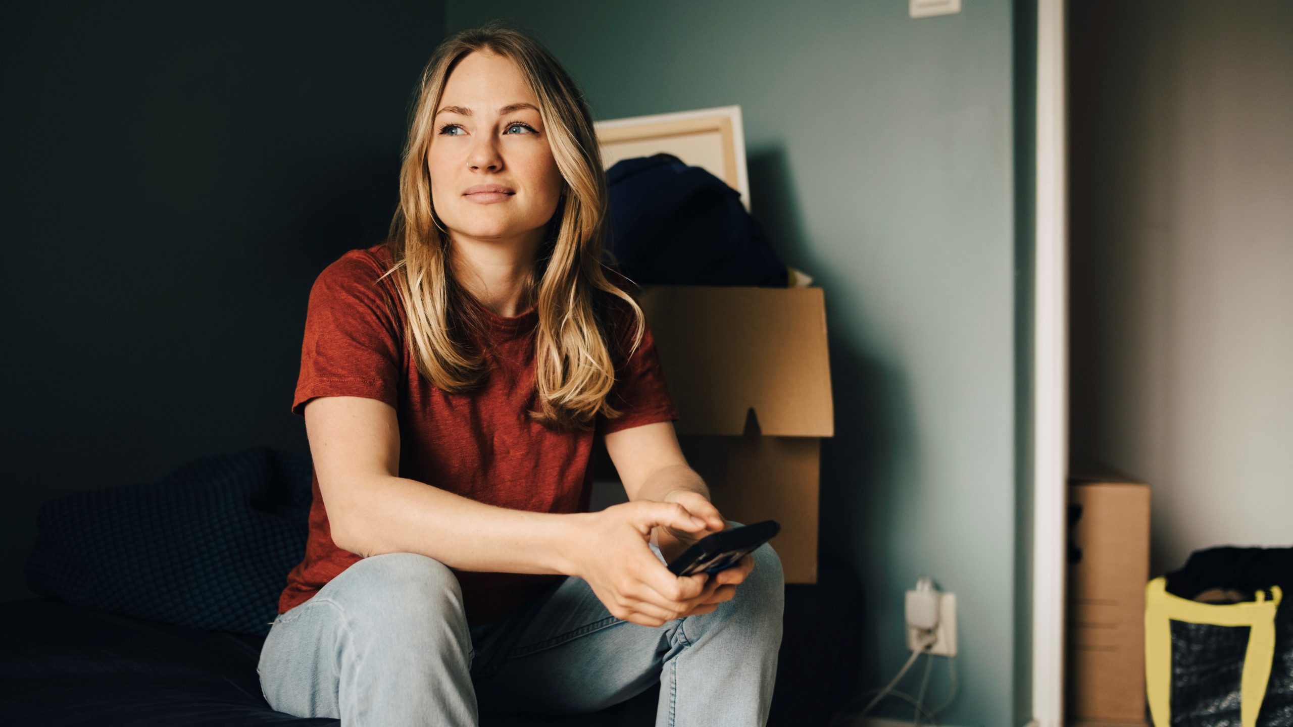 Thoughtful young woman looking away while sitting with smart phone in bedroom