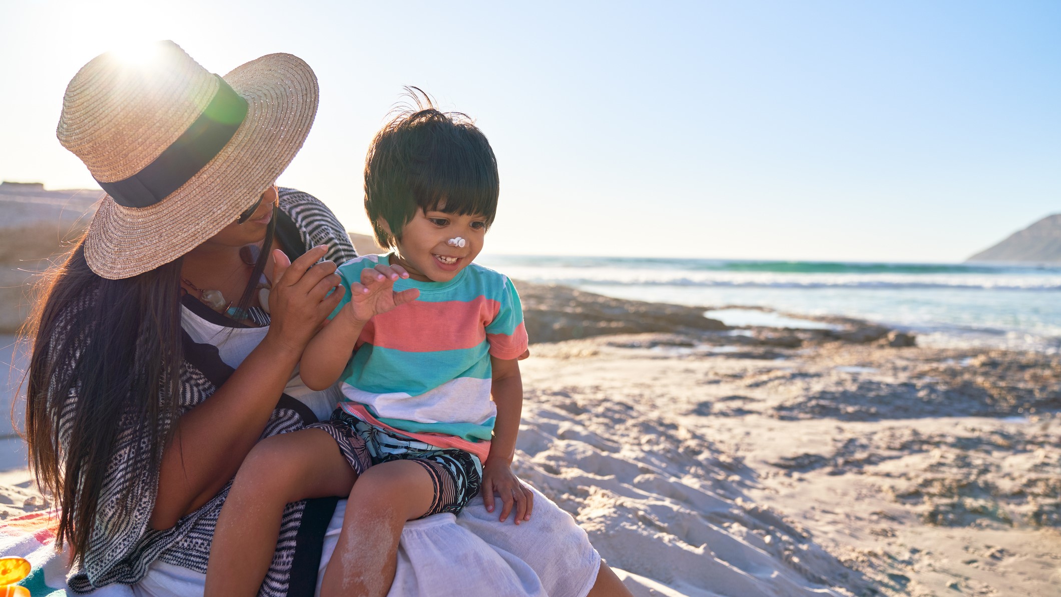 Mother applying sunscreen to nose of cute son on sunny beach, enjoying summer vacation.