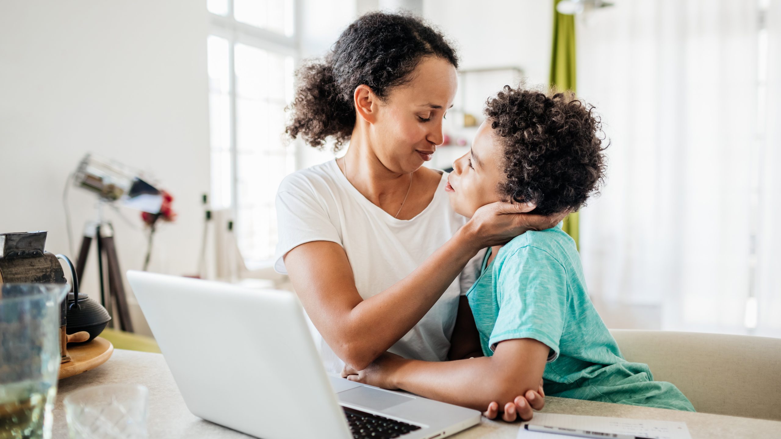 A single mom being affectionate with her young son while working from home on her laptop.